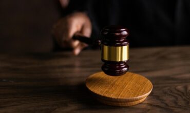 A close-up photograph of a wooden judge's gavel resting on a sounding block on a dark wooden table, representing the legal system and judicial rulings.